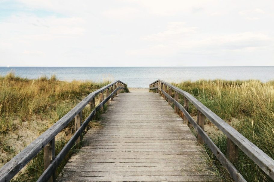 Holzsteg führt durch Graslandschaft zum Meer unter bewölktem Himmel.