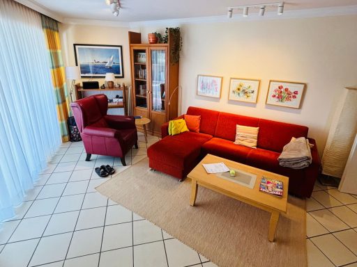 Wohnzimmer mit rotem Sofa, Lesesessel, Tisch und Regalen, helle Farbgestaltung.