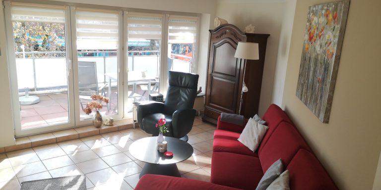 Heller Wohnraum mit rotem Sofa, Sessel, Tisch und Fenster mit Blick ins Freie.