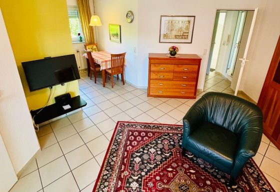 Ein Wohnzimmer mit grünem Sessel, TV, Essbereich und Kommode auf gefliestem Boden.