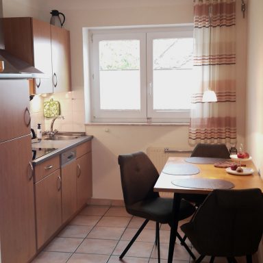 Helle Küche mit Holzschränken, Tisch und Stühlen, Fenster mit Vorhängen.