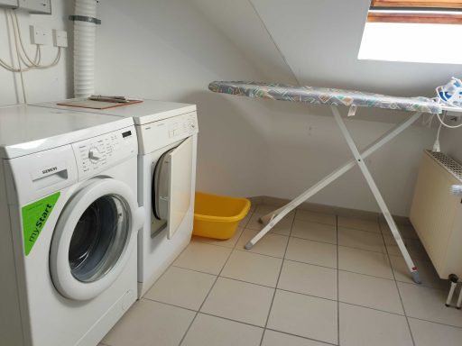 Waschmaschine und Trockner neben einem Bügelbrett in einem hellen, neutralen Raum.