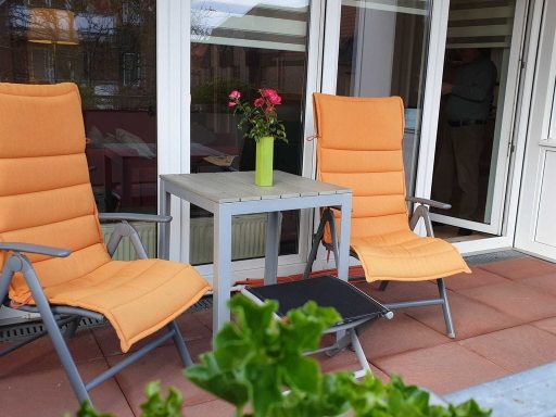 Zwei orangefarbene Liegestühle neben einem kleinen Tisch mit einer Vase und Rosen.